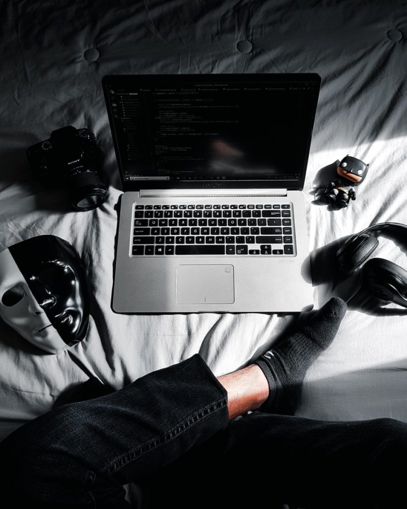 MacBook in middle of camera, action camera, mask, and headphones on bed